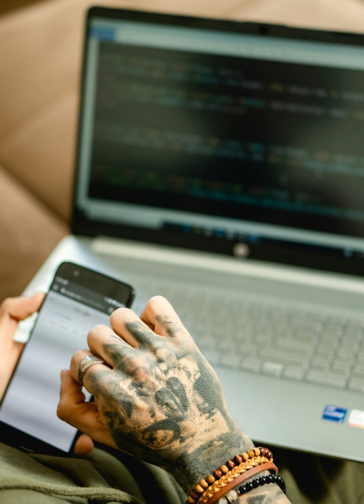 Close-up of a tattooed hand holding a smartphone, working on a laptop in an office setting.